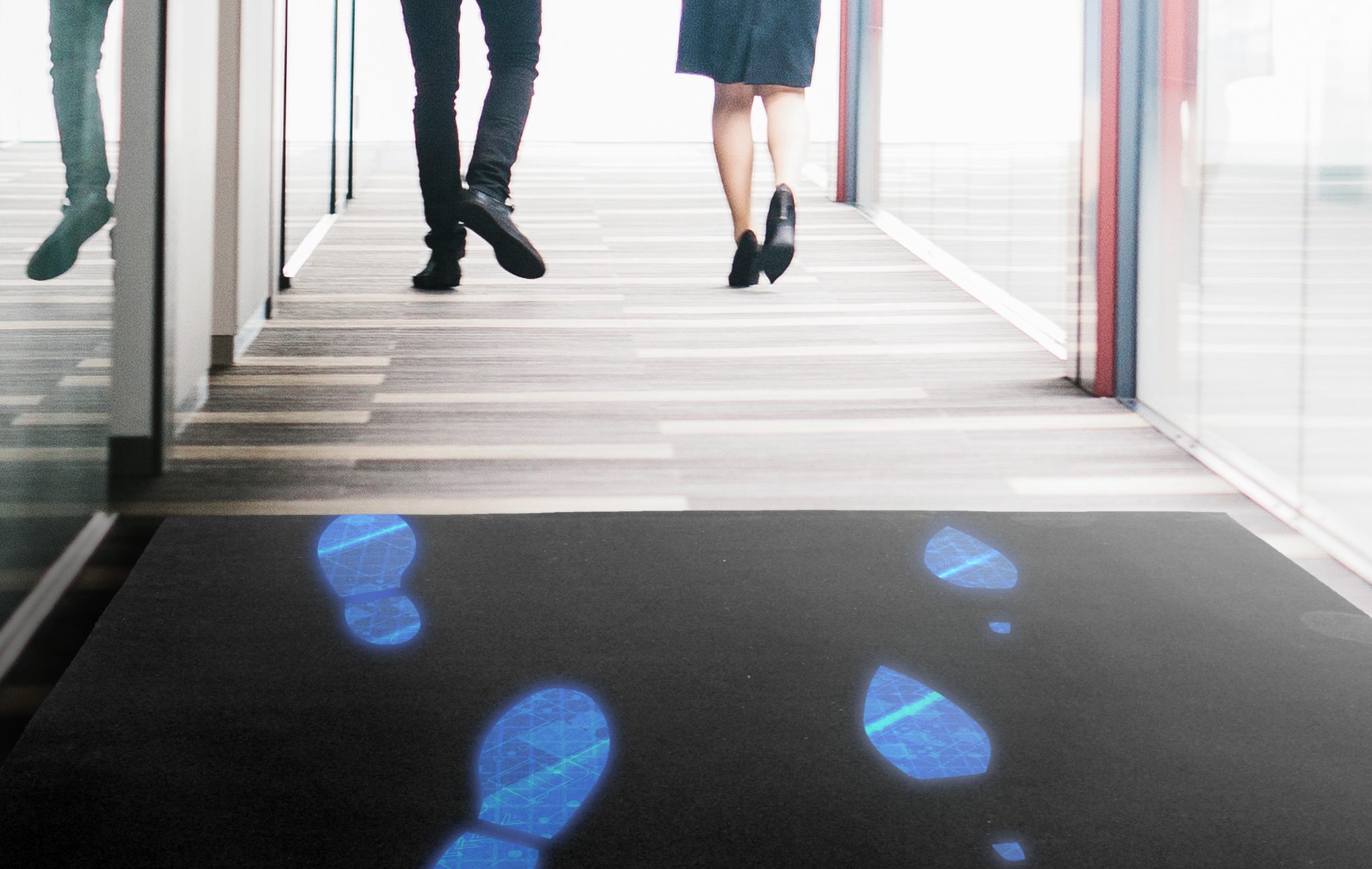 Ein Mann und eine Frau (man sieht nur die Beine) gehen einen Gang entlang. Auf dem schwarzen Teppich leuchten ihre Fussabdrücke blau auf.