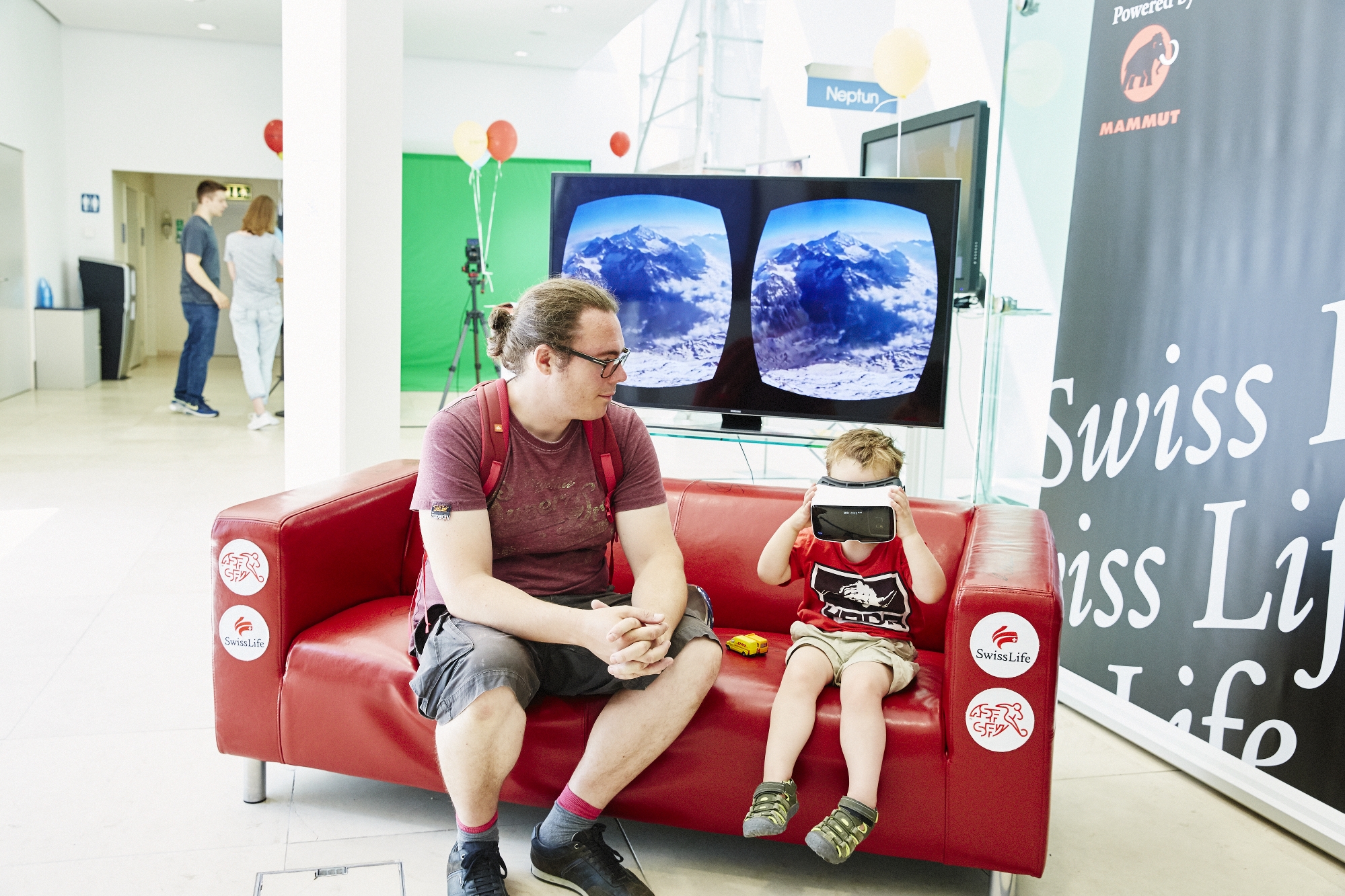 Ein Mann sitzt mit einem Kleinkind auf einem roten Sofa. Das Kind hält eine VR-Brille vor seine Augen.