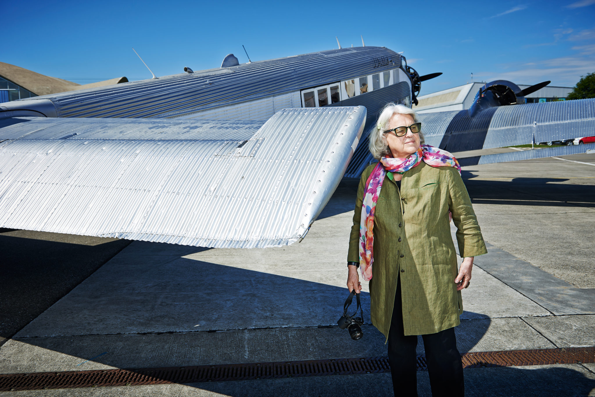 Eine ältere Dame mit auffällig dicker Brille und einem bunten Schal steht vor einem silbernen Flugzeug und schaut nach rechts.