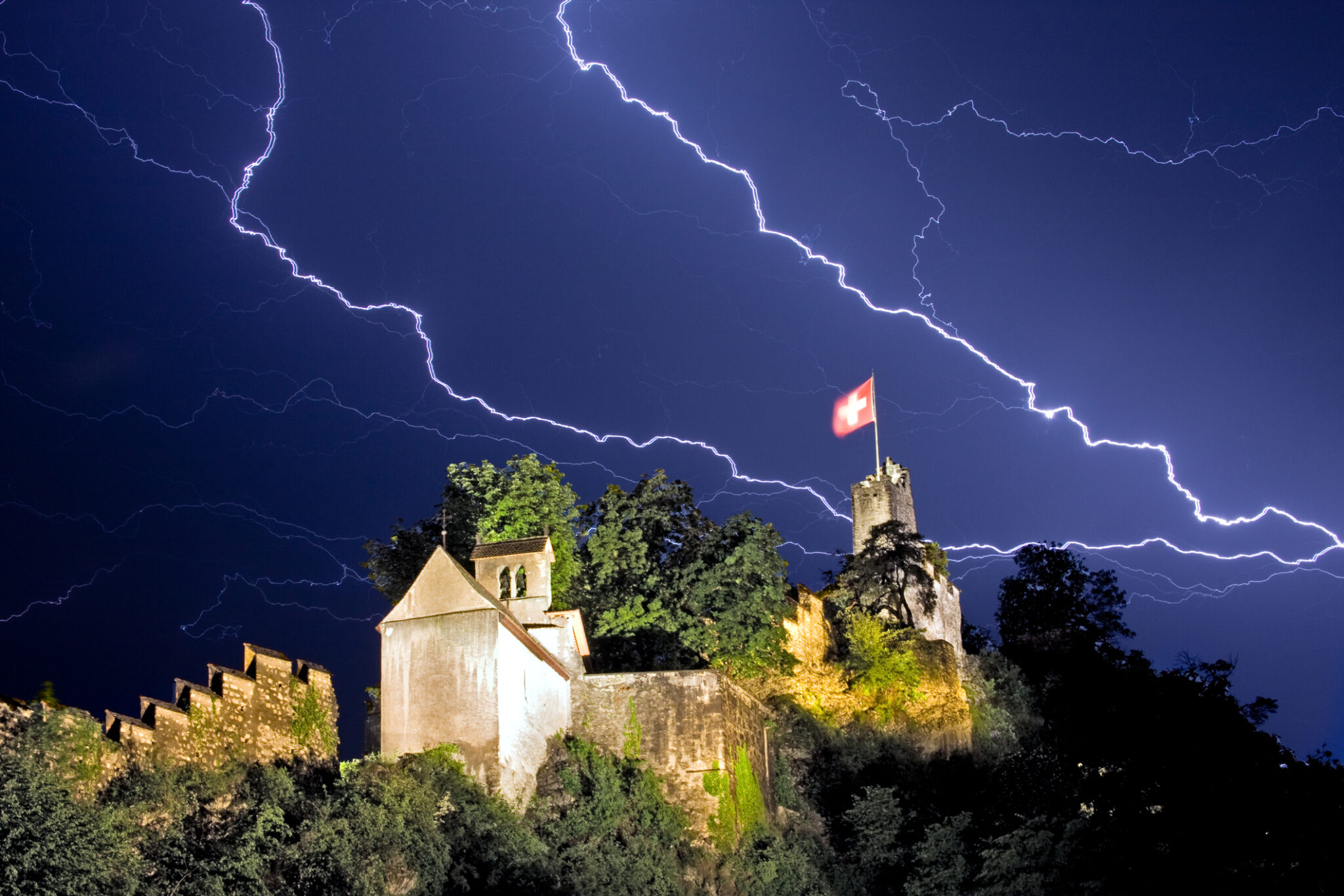 A bolt of lightning crosses the sky over the Stein castle in Baden Switzerland. A Swiss flag is shown waving in the forceful wind over the castle.