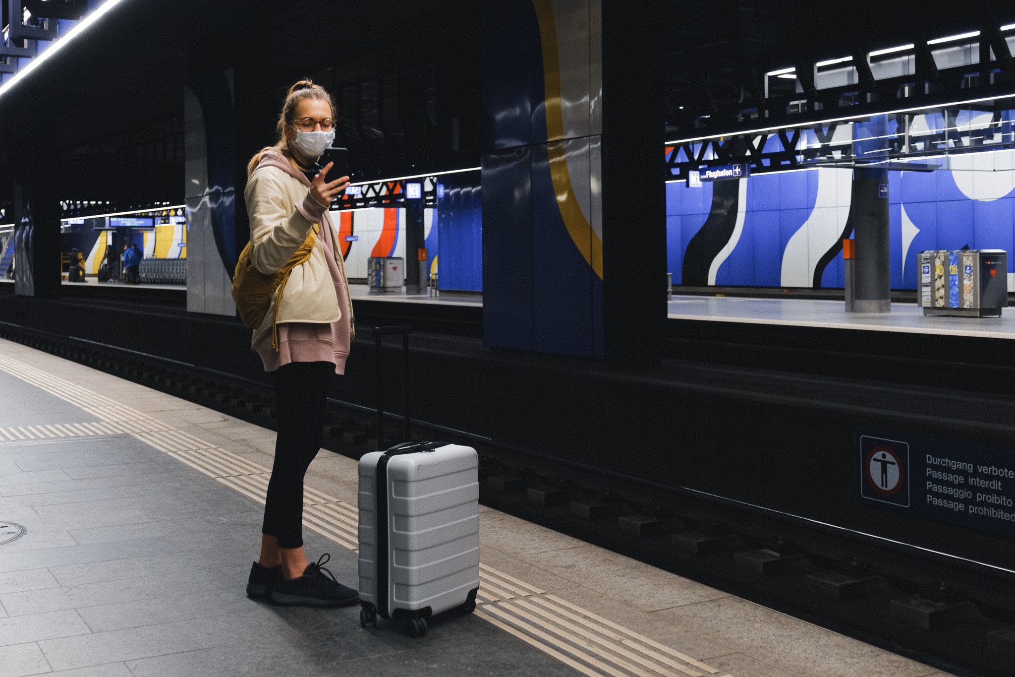 Frau mit Schutzmaske steht auf einem Bahnsteig und schaut auf ihr Handy,