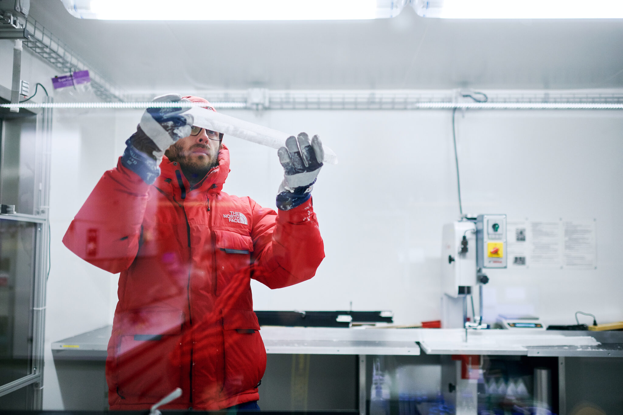 Ein Forscher in roter Winterjacke und blau-weiss gestreiften Handschuhen hält einen Eisbohrkernen in den Händen.