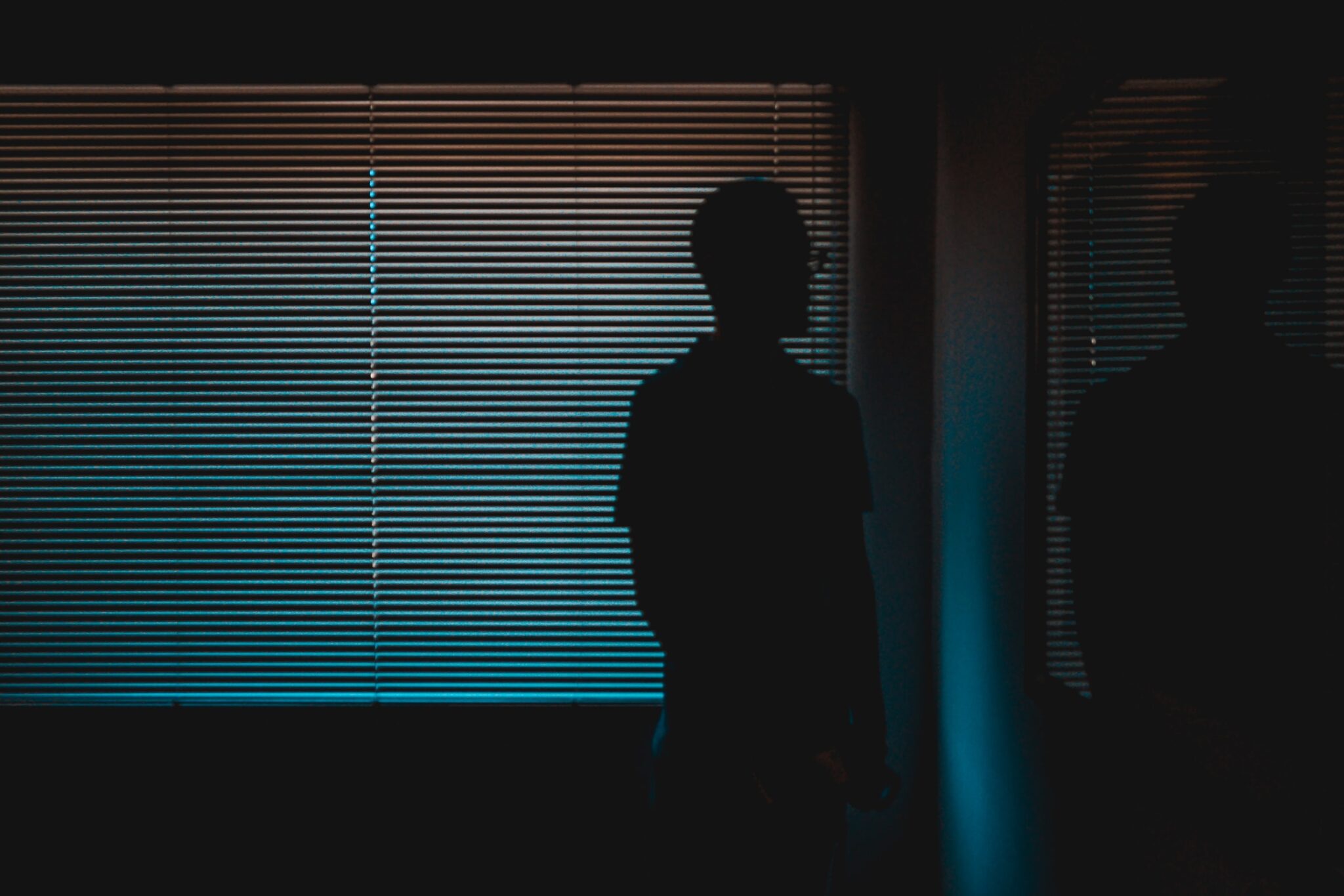 Ein Mann steht in einem abgedunkelten Raum vor heruntergelassenen Jalousien, durch die ein wenig Licht in blau und orange fällt. Man sieht nur seinen Schattenriss.