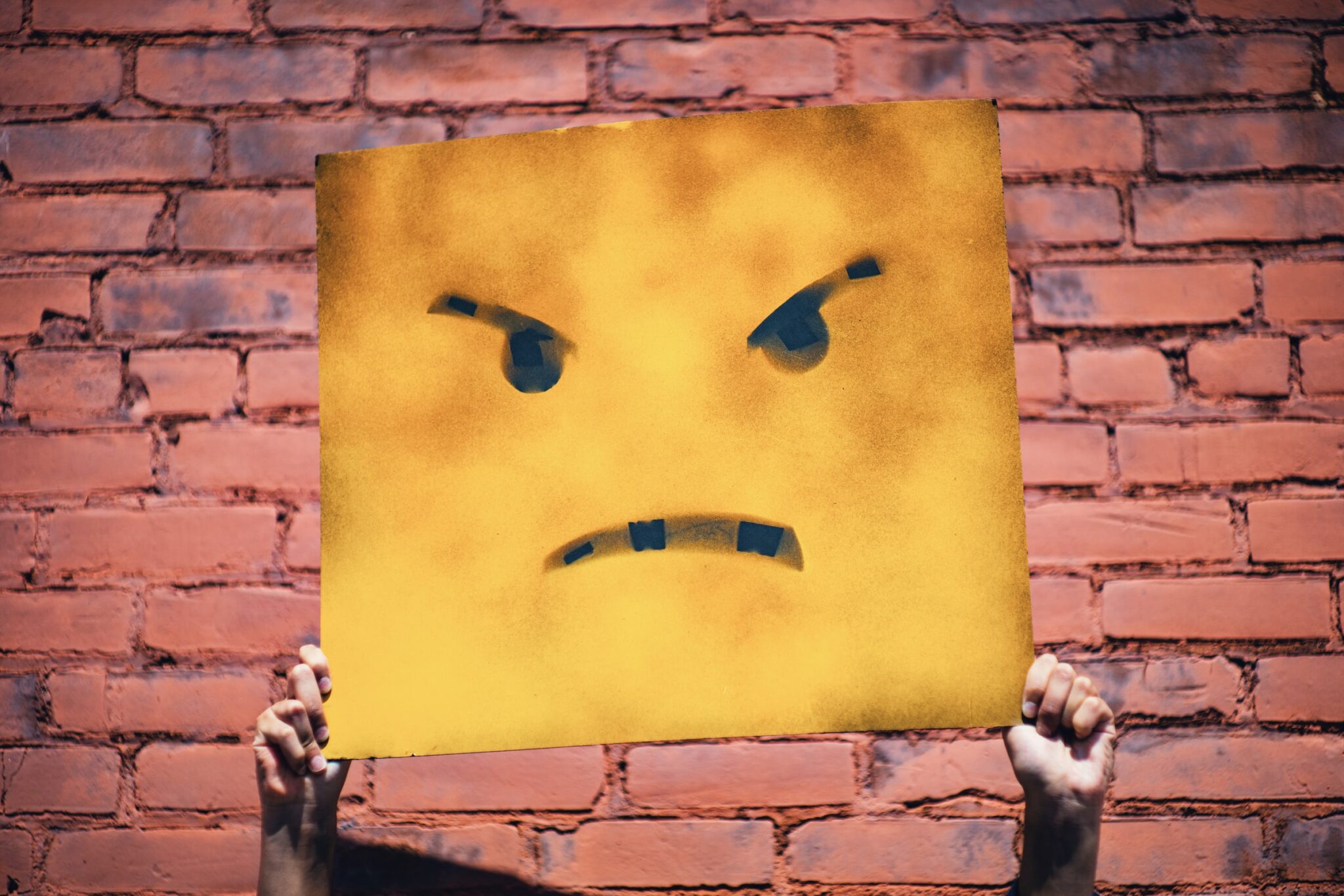 Ein wütendes Emoji auf gelbem Karten wird vor einer Backsteinwand hochgehalten.