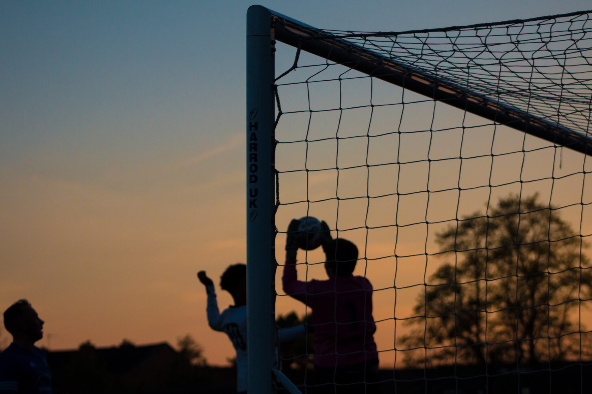 Fussballspielende Menschen als Schattenriss im Sonnenuntergang halb durch ein Fussballtornetz fotografiert.