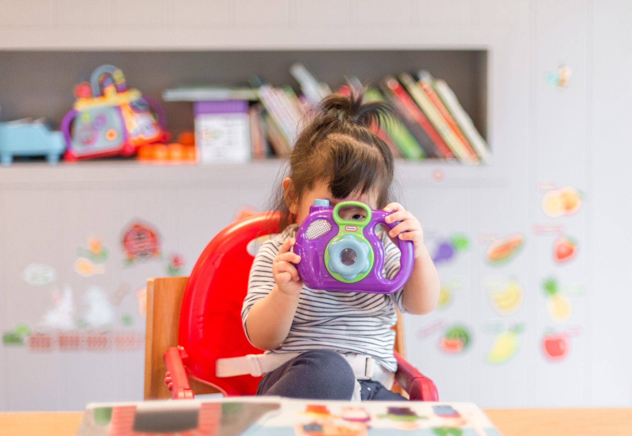Ein kleines Mädchen sitzt in einem Hochstuhl und schaut durch eine lila Spielzeugkamera.