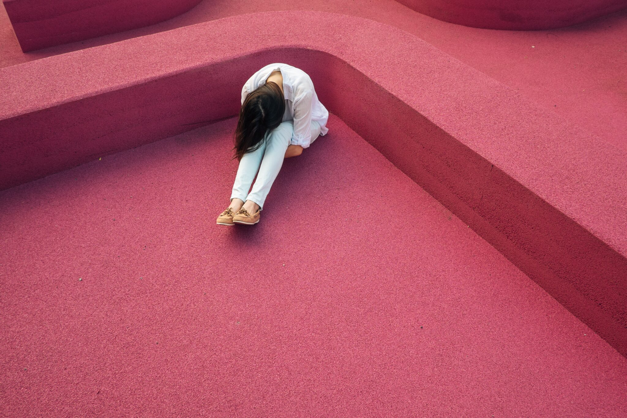 Eine Frau sitzt auf rotem Untergrund alleine und erschöpft in einer Ecke und legt den Kopf auf ihre angezogenen Knie.