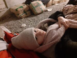 Ein Selfie zeigt Oksana Hela wie sie mit Mütze und Schal auf dem Boden im Keller schlafen muss. Im Hintergrund sind wild Kartonboxen zu erkennen.