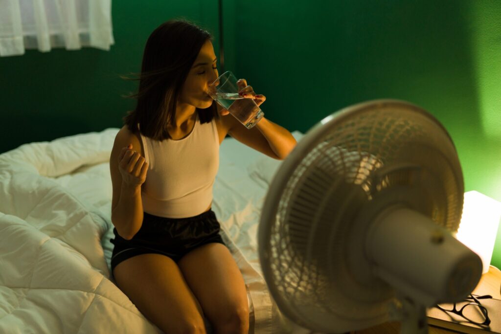 Eine Frau sitzt auf dem Bett vor einem Ventilator und trinkt ein Glas Wasser.